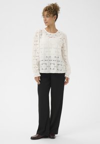 Žena s kovrčavom kosom u punđi, koja nosi bijelu čipkastu bluzu s dugim rukavima, crne široke hlače i tamnosmeđe cipele, stoji na bijeloj pozadini.