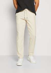 Pantaloni leggeri beige con vita regolabile, gambe affusolate e una texture sottile. Abbinati a sneaker bianche.