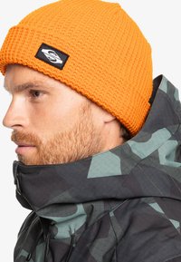 Oranžová pletená čepice s texturovaným vzorem a černou nášivkou loga. Nosená s tmavou kramkovou bundou. Detailní profilový pohled.