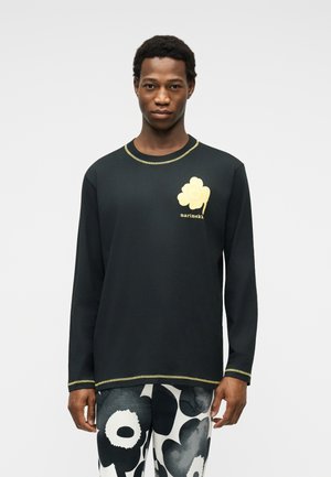 Man wearing black long-sleeve shirt with yellow flower logo and "marimekko" text, paired with black-and-white floral patterned pants.