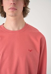 Long-sleeve shirt in coral cotton, featuring a round neckline and a small red logo on the chest. Smooth texture, casual fit.