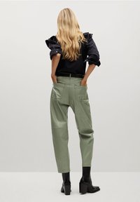 Top negro con mangas abullonadas, pantalones de imitación piel verdes con bolsillos laterales, botas negras de tacón alto. Toma desde la parte trasera contra un fondo neutro.
