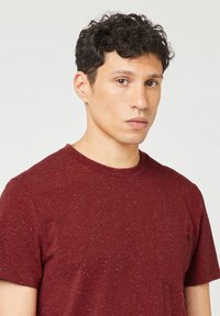 Jonge man met kort, krullend donker haar, gekleed in een gespikkelde maroon T-shirt, die iets opzij kijkt tegen een effen achtergrond.