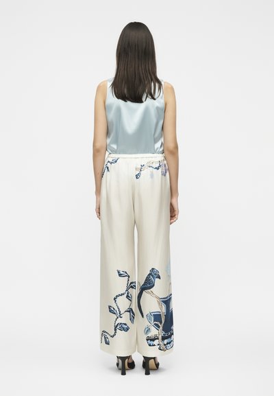 Femme portant un haut sans manches en satin bleu clair et un pantalon large crème avec des motifs d'oiseaux bleus et de vignes, debout de dos.