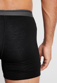 Közelkép egy személy alsó törzséről és a felső combjáról, aki fekete boxer alsóneműt visel szürke derékpánttal, a keze nyugodtan a teste mellett.