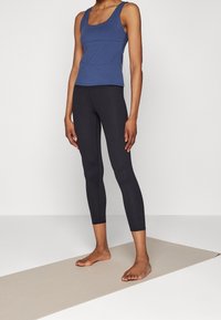 Osoba stoi boso na beżowej macie do jogi, ubrana w czarne legginsy i niebieską bluzkę bez rękawów, na jednolitym tle.