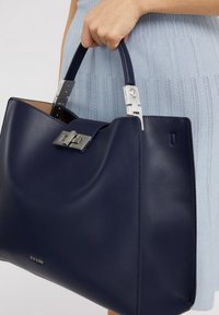 Granatowa skórzana torebka o gładkiej fakturze, z srebrnymi okuciami, zapięciem na klamrę i solidnymi uchwytami, trzymana w jednej dłoni.