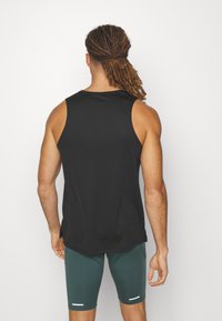 Homme avec des dreadlocks portant un débardeur noir et un short de sport vert foncé, debout face à un mur blanc.
