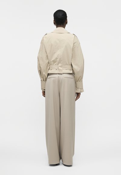 Άτομο με σκουρόχρωμο δέρμα και μαλλιά δεμένα πίσω που φοράει μπεζ κοντό σακάκι με ζώνη στη μέση και μπεζ φαρδιά παντελόνια, όρθιο με πλάτη.