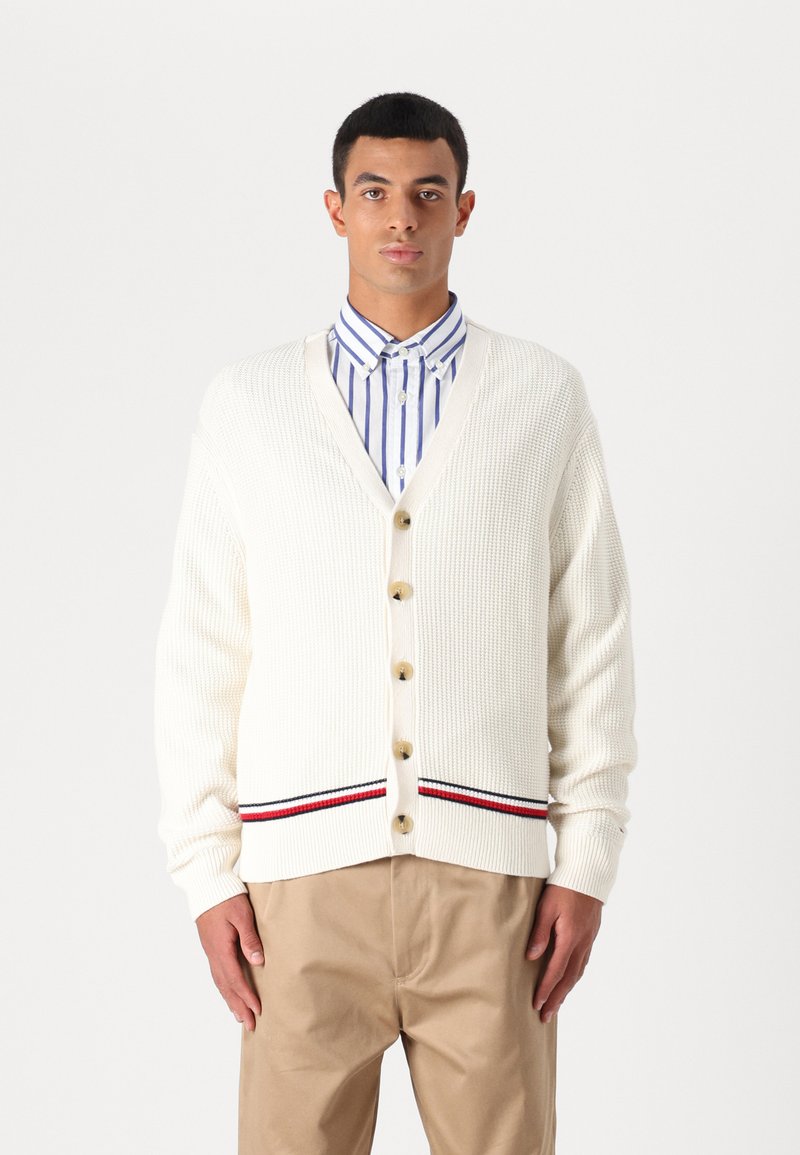 Kremfarget cardigan med strukturert strikk, med seks store knapper og et stripete detaljer i rød og marineblå ved kanten.