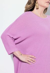 Übergroßer rosa Strickpullover mit gerippter Textur, weiten 3/4-Ärmeln und einem Rundhalsausschnitt. Mit metallischen Akzenten in den Ohrringen.