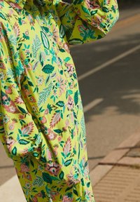 Robe florale jaune vif avec des motifs botaniques verts et roses, manches longues et coupe ample, avec des fermetures à boutons et un col.
