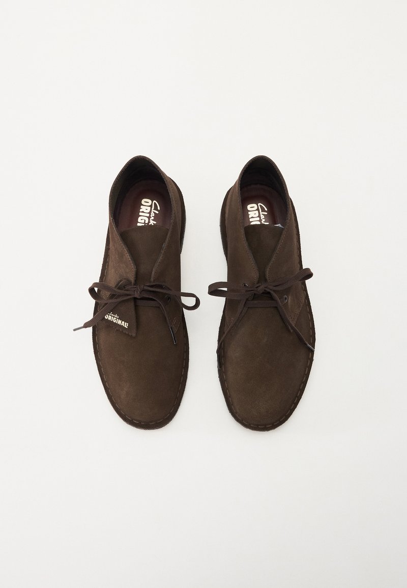 Bruine suède schoenen met een afgeronde neus, vetersluiting en gestikte details. Bevat een subtiele label met "Clark Originals." Soepele textuur.