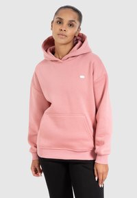 Hoodie rose clair avec une poche avant, capuche à cordon, et un petit logo ovale sur la poitrine. Tissu doux et coupe décontractée.