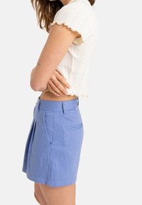 Bermuda azzurra chiara, sartoriale, in tessuto texturizzato, con tasche laterali e pieghe frontali, abbinata a una blusa bianca a maniche corte.