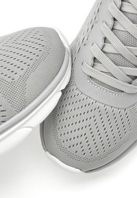 Graue Sportschuhe mit einem oberen Teil aus Netzmaterial und durchbrochenem Design, ausgestattet mit flachen Schnürsenkeln, einem gepolsterten Schaft und einer weißen Sohle.