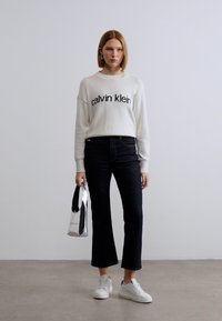 Λευκό πουλόβερ με μαύρο κείμενο «calvin klein», σκούρο τζιν με φουσκωτά μπατζάκια, λευκά αθλητικά παπούτσια και ασημί τσάντα χειρός στο αριστερό χέρι.