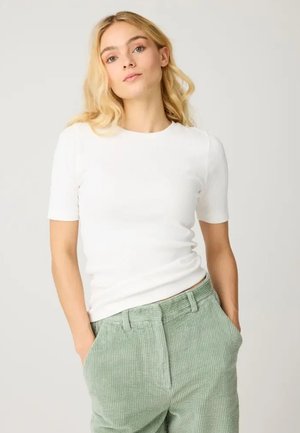 Donna bionda che indossa una maglietta bianca a maniche corte e pantaloni di velluto a coste verde chiaro, con le mani nelle tasche, in piedi davanti a uno sfondo semplice.