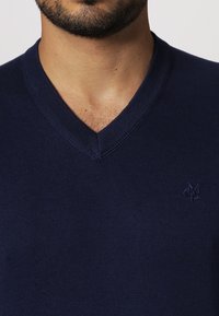 Ciemnoniebieski sweter z dzianiny z dekoltem w serek i prążkowanymi detalami, z małym haftowanym logo po lewej stronie.