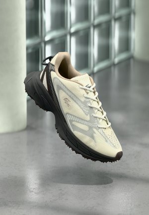 Scarpa da ginnastica beige chiaro e grigia con suola nera sospesa a mezz'aria contro una parete piastrellata e uno sfondo di pavimento grigio.