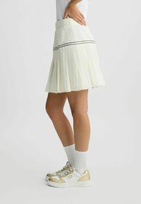 Faltenrock in Weiß mit einem strukturierten, kontrastierenden Bund und schwarzem Akzentstreifen. Kombiniert mit weißen Sneakers mit goldenen Akzenten und weißen Socken.