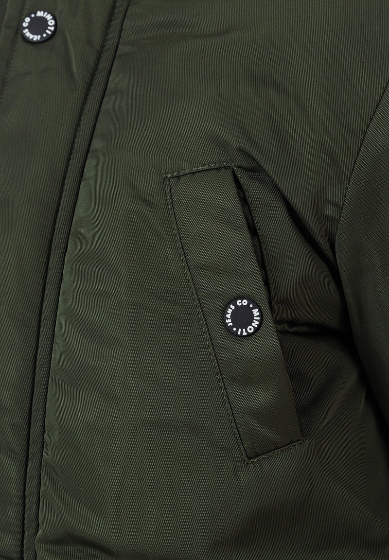 Chaqueta acolchada verde oscuro con un bolsillo con cremallera y cierre de botón negro, que muestra un patrón de tejido texturizado.