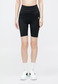 Fekete kerékpáros rövidnadrág magas derékrésszel és logóval díszítve. Sima, rugalmas anyag. Fehér sportcipővel és zoknival párosítva.