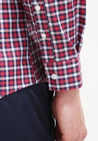 Rood en marineblauwe geruite blouse met lange mouwen, voorzien van witte knopen en een knoopmanchet. De stof heeft een gladde textuur met een klassiek ruitpatroon.