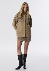 Tan suède jacket met decoratieve borduursels en studs, gecombineerd met een bijpassende minirok met vergelijkbare details. Zwarte laarzen tot de knie.