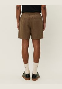 Pantalones cortos marrones hasta la rodilla con una cinturilla elástica y un bolsillo trasero, hechos de una tela texturizada. Llevados con zapatos negros y calcetas claras.
