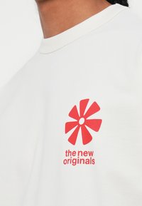 Balta medvilninė marškinė su raudonu gėlių grafiku ir tekstu "nauji originalai" po juo. Apvalus kaklas ir trumpi rankoviai.