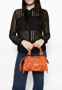 Borsa a mano in pelle arancione con molteplici tasche, dettagli con borchie e frange; tenuta da una persona che indossa una blusa trasparente nera a righe.