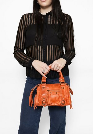 Oranje leren handtas met meerdere zakken, studaccenten en franjes; vastgehouden door een persoon die een zwart, gestreept doorschijnend blouse draagt.