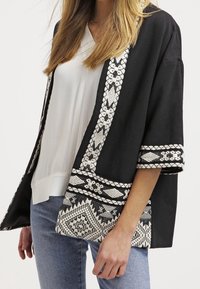 Kvinde iført en sort kimono-stil jakke med hvid geometrisk broderi over en hvid bluse og blå jeans, skåret ved midtsektionen.