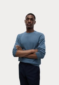 Hellblauer Strickpullover mit Rundhalsausschnitt, gerippten Bündchen und detaillierten Nähten. Das Modell trägt eine marineblaue Hose, die Arme verschränkt, vor einem neutralen Hintergrund.