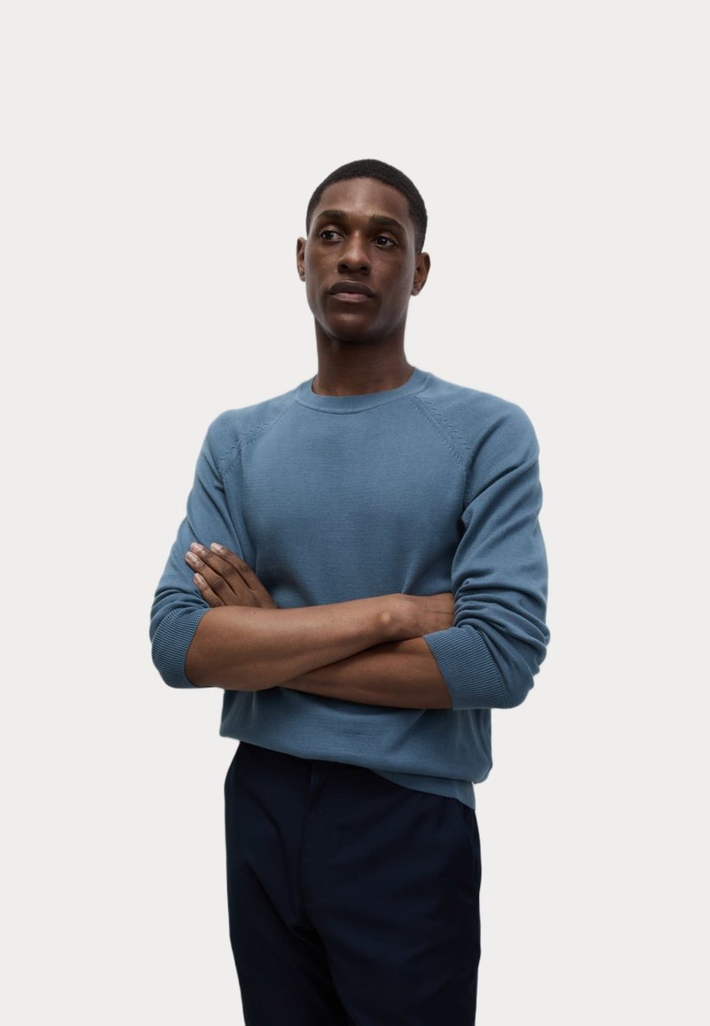 Hellblauer Strickpullover mit Rundhalsausschnitt, gerippten Bündchen und detaillierten Nähten. Das Modell trägt eine marineblaue Hose, die Arme verschränkt, vor einem neutralen Hintergrund.