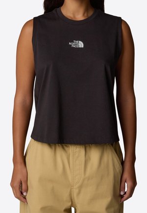 Woman wearing a sleeveless black cropped top with a white "The North Face" logo and beige high-waisted pants, hands in pockets.
