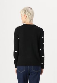 Pull noir à manches longues, avec des accents en pompons blancs sur les manches. Fabriqué dans un matériau doux et texturé. Encolure ras du cou.
