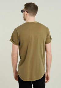 T-shirt verde oliva a maniche corte con risvolti, scollatura rotonda, orlo leggermente curvato e una finitura morbida e texturizzata. Pantaloni neri e occhiali da sole.