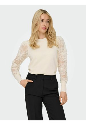 Pull crème avec des manches en dentelle transparente et texturée. Le buste est uni, complété par un pantalon noir ajusté. Manches à bords ribbés simples.