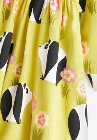 Grünes Baumwollkleid mit schwarz-weißen Panda-Illustrationen und rosa Blumenmustern vor einem grasbewachsenen Hintergrund.