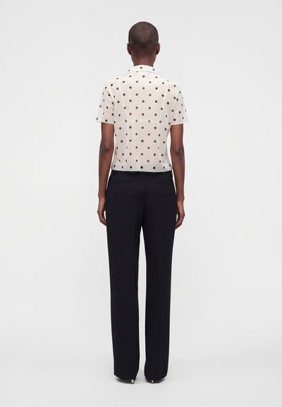 Personne portant une chemise blanche à manches courtes avec des pois noirs et un pantalon droit noir, debout de dos sur un fond clair uni.