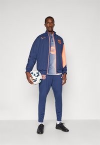 Marineblå atletisk træningsdragt med orange detaljer, bestående af en lynlåsjakke og stramme bukser. Person holder en fodbold og har sorte sko på.