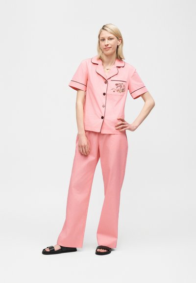 Frau steht mit einer Hand in der Hüfte, trägt ein rosa Kurzarm-Pyjama-Set mit bestickter Brusttasche und schwarze Pantoletten vor weißem Hintergrund.