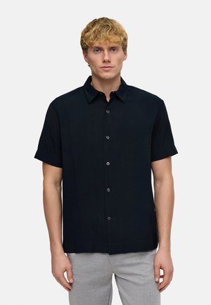 Jeune homme aux cheveux blonds courts portant une chemise noire à manches courtes et un pantalon gris clair sur un fond uni.