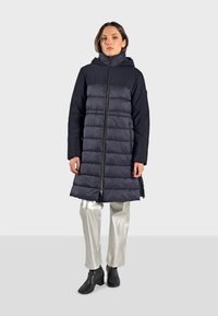 Abrigo acolchado navy con cierre frontal, gran capucha y diseño de paneles. Combinado con pantalones plateados brillantes y botas negras de tobillo. Textura de tela suave.