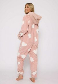 Lyserød fleece-onesie med hvide hjerte mønstre, hætte og elastiske manchetter. Blød tekstur og afslappet pasform, velegnet til hjemmebrug.