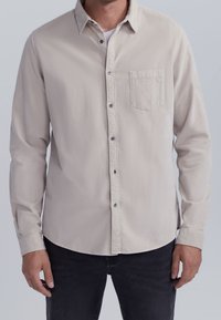 Camisa beige clara de manga larga con botones, con un bolsillo en el pecho, hecha de una tela suave, con un cuello clásico y botones oscuros.