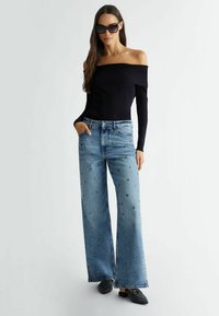 Melna off-shoulder ribbed blūze, apvienota ar gaiši ziliem platām biksēm ar sudraba dimanta formas rotājumiem. Valkā melnus loaferu tipa apavus.