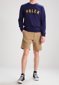 Sweatshirt bleu marine avec le texte "POLER" en jaune, assorti à un short kaki. Le modèle porte des baskets noires et blanches. Coupe décontractée et confortable.